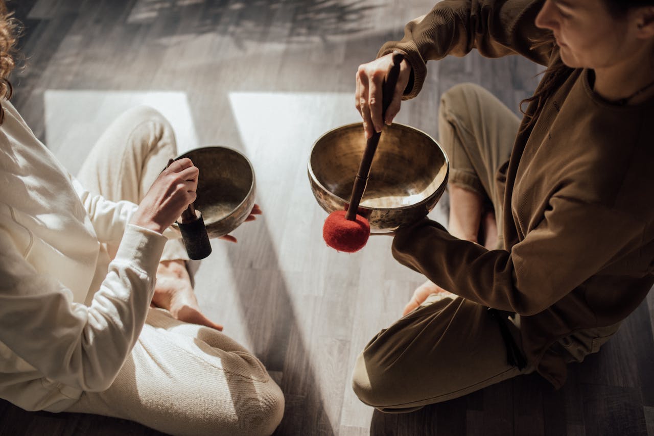 Two women meditating with Tibetan singing bowls in a peaceful indoor setting.